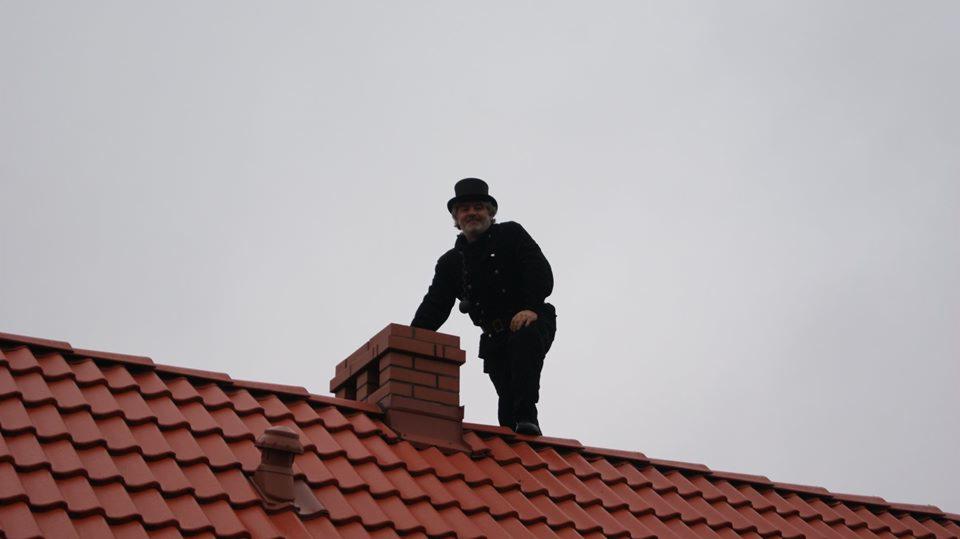Kominiarz w tradycyjnym stroju (czarny frak i cylinder) stoi na dachu z czerwonej dachówki obok ceglanego komina, z widoczną szczotką kominiarską.