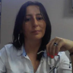 Portret kobiety w białej koszuli z okularami w dłoni, na neutralnym tle.