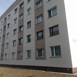 Wielopiętrowy budynek po renowacji elewacji, z dwukolorową fasadą (beż i brąz), widoczny znak parkingu dla niepełnosprawnych i ulica F.S. Składkowskiego na bocznej ścianie.