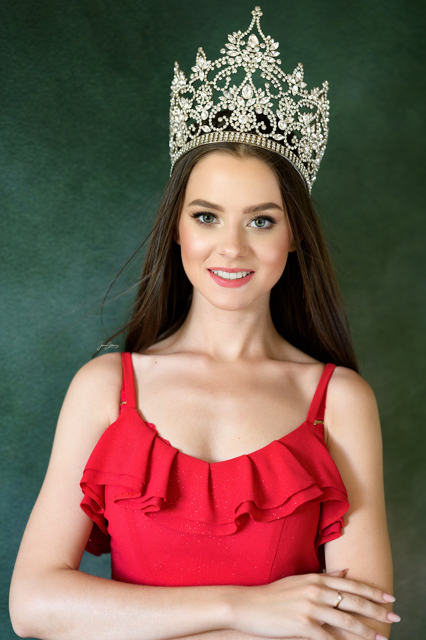 Portret uśmiechniętej młodej kobiety w czerwonej sukience z falbanami i bogato zdobionej koronie na głowie, wykonany w Szczecinie na tle ciemnozielonej ściany.