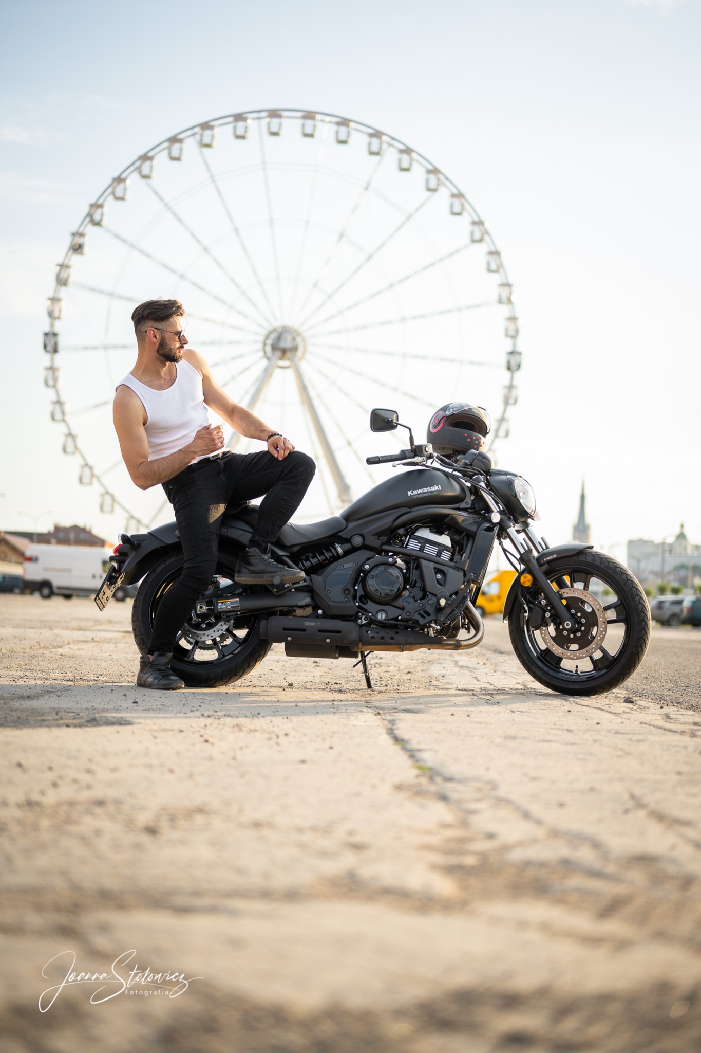 Mężczyzna w białym podkoszulku i czarnych spodniach pozuje siedząc na czarnym motocyklu Kawasaki na tle diabelskiego młyna i miejskiego krajobrazu.