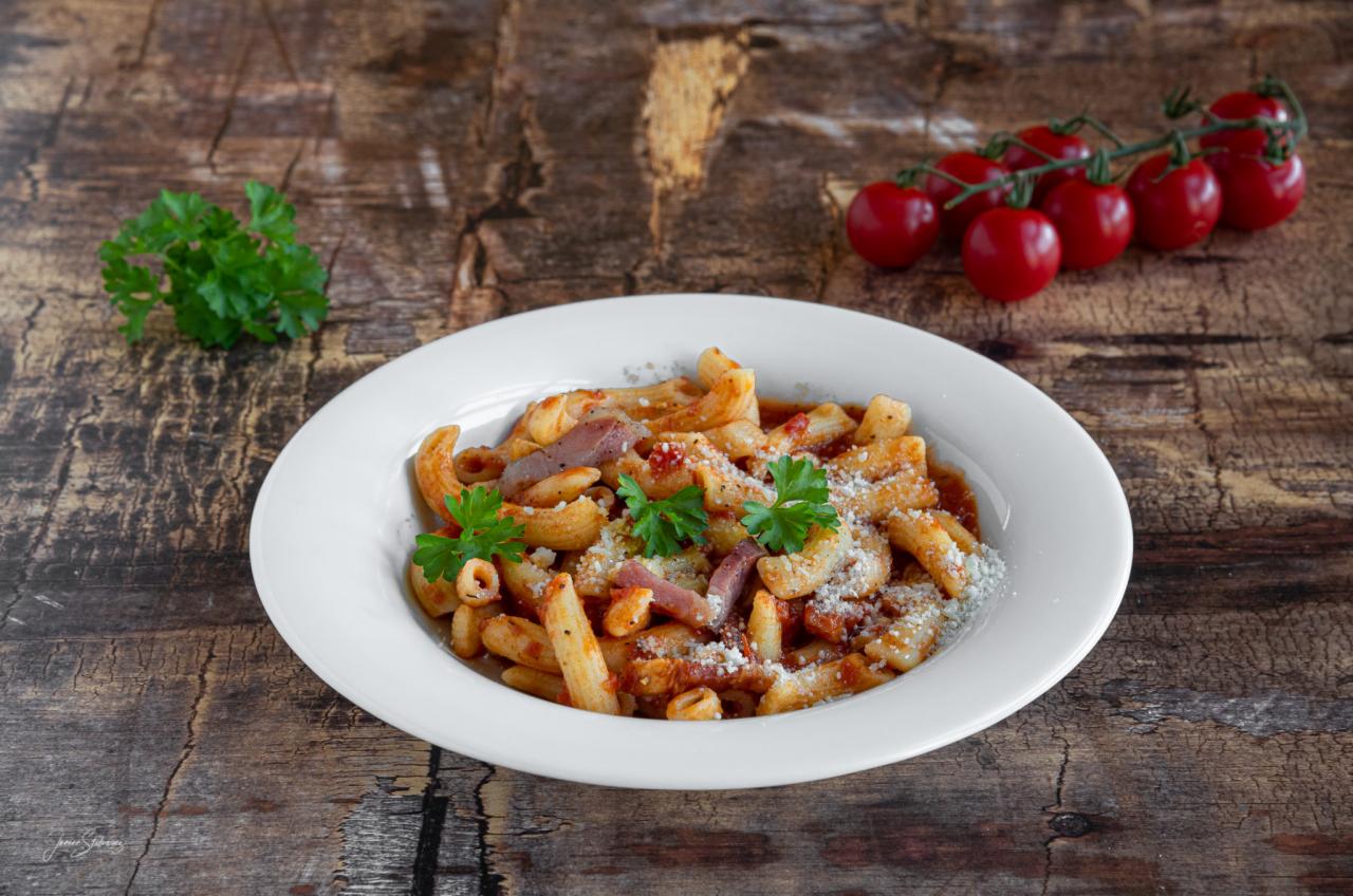 Makaron casarecce w sosie pomidorowym z szynką parmeńską i posypką serową, udekorowany świeżą natką pietruszki, podany na białym talerzu na drewnianym stole, z gałązką pomidorków cherry i kępką...