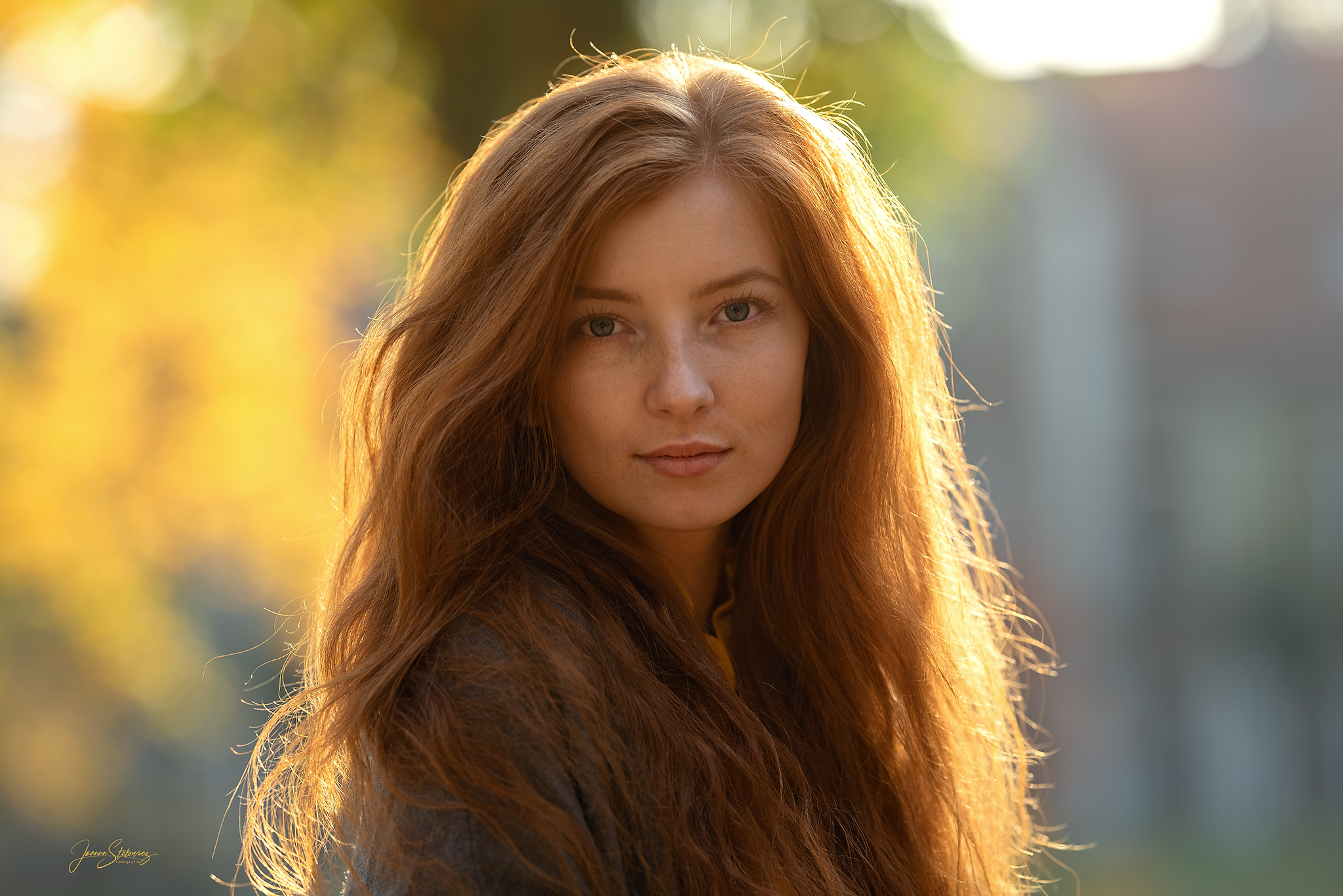 Portret kobiety o długich, rudych włosach, z delikatnym makijażem i jasnymi oczami, na rozmytym tle jesiennych drzew w ciepłym, naturalnym świetle.