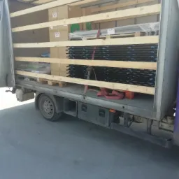 Wnętrze otwartej ciężarówki z ładunkiem zabezpieczonym drewnianymi belkami: palety z kartonami, czarne metalowe kraty w folii i czerwone pasy mocujące.