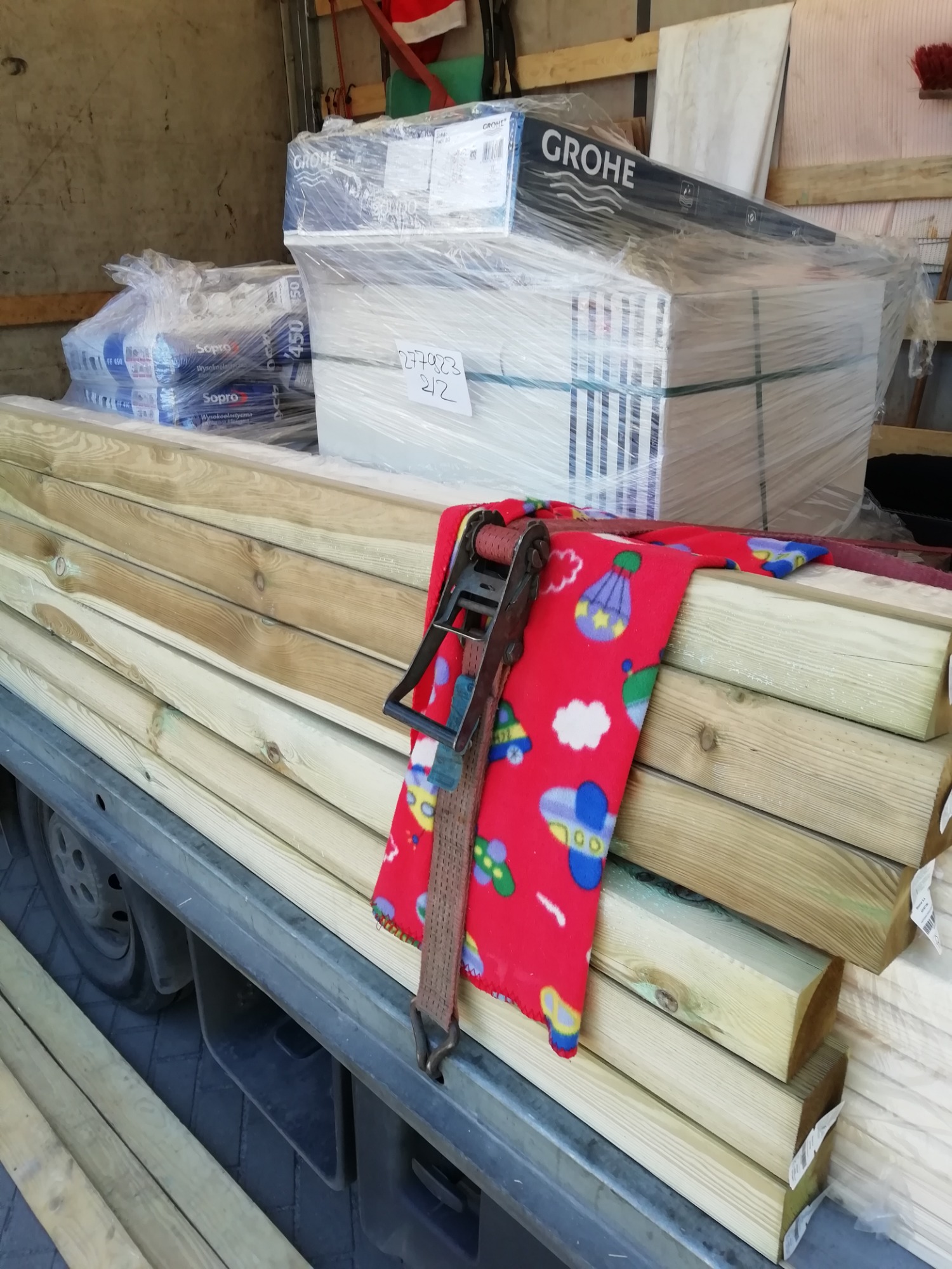 Załadunek materiałów budowlanych: kantówki, płytki, zaprawa Sopro i armatura Grohe zabezpieczone pasem transportowym wewnątrz pojazdu.