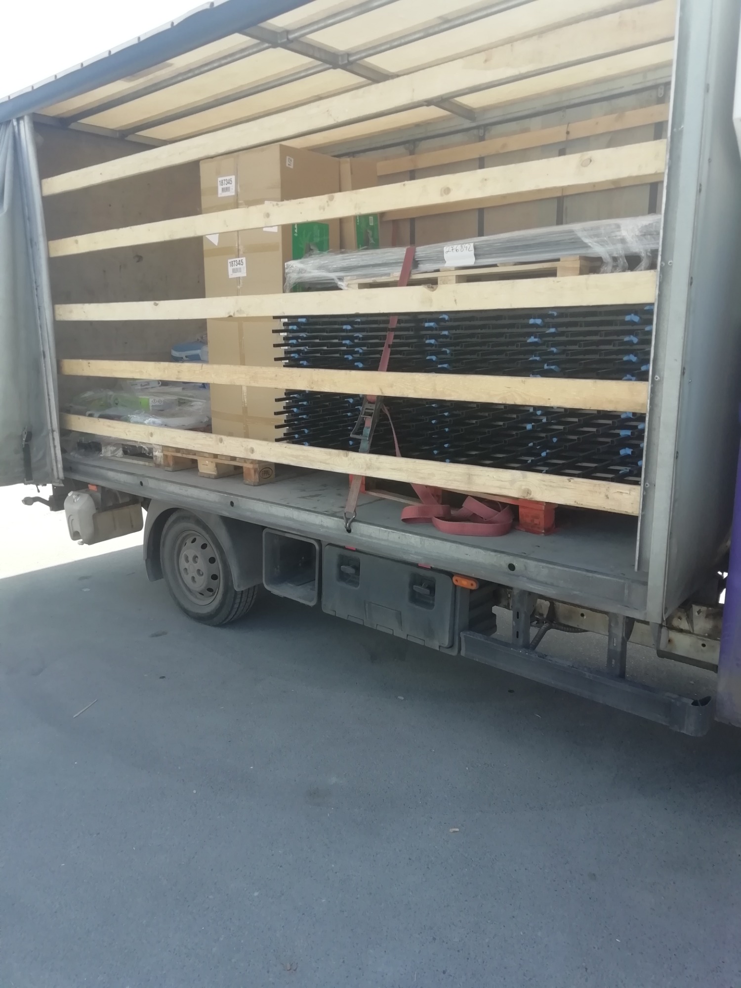 Wnętrze otwartej ciężarówki z ładunkiem zabezpieczonym drewnianymi belkami: palety z kartonami, czarne metalowe kraty w folii i czerwone pasy mocujące.