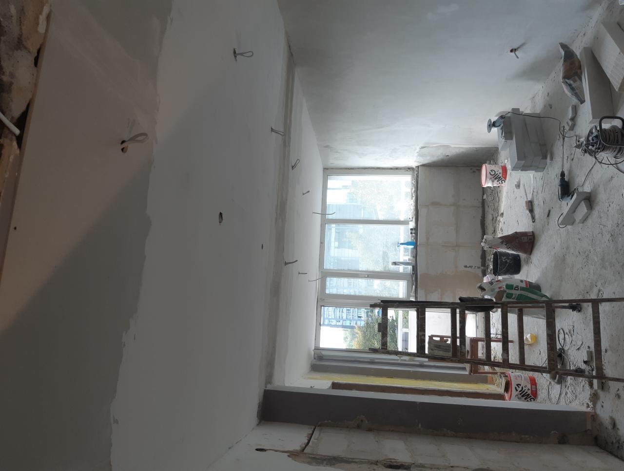 Wnętrze w trakcie prac przygotowawczych do wykończenia, widoczne częściowo wyszpachlowane ściany, okno, drabina, narzędzia i materiały budowlane.