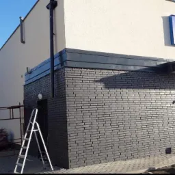 Wykończona elewacja budynku z szarej cegły klinkierowej, z widocznym fragmentem białej elewacji, stalową listwą okapową i fragmentem okien oraz wejścia do budynku, drabina i rusztowanie w tle.