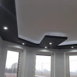 Nowoczesny sufit podwieszany z czarnymi elementami i punktowym oświetleniem LED, widok z dołu, trzy okna z widokiem na pole w tle.