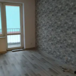 Wykończone wnętrze pokoju z drewnopodobną podłogą, ścianą z dekoracyjnej cegły w odcieniach szarości i białym oknem balkonowym.