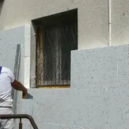 Pracownik w białym kombinezonie i kasku, z widocznym logotypem na szelkach, aplikuje materiał izolacyjny na ścianę budynku obok okna z kratą, używając szpachelki.