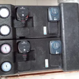 Dwa moduły rozdzielacza z pompami obiegowymi i manometrami, podłączone do instalacji z rur miedzianych, zamontowane na ścianie z płytek.