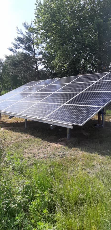 Panele fotowoltaiczne zamontowane na metalowej konstrukcji na trawiastym terenie, widoczne drzewa w tle, częściowo zasłonięta osoba pracująca przy instalacji.