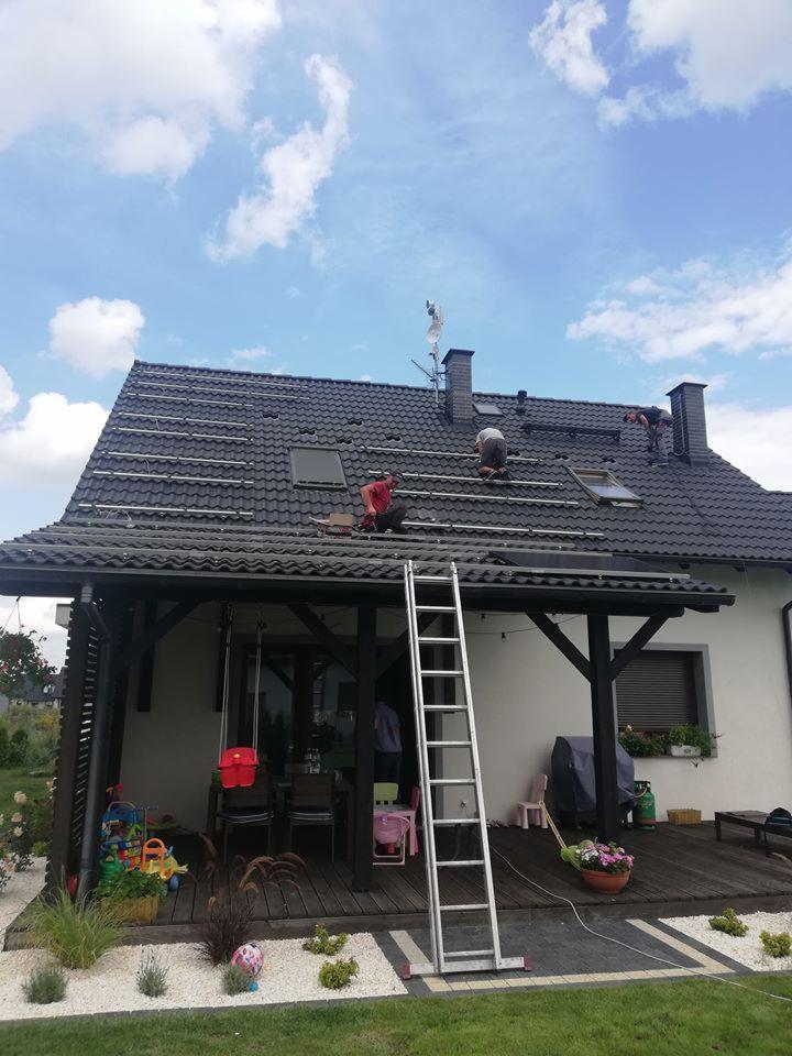 Montaż paneli na dachu domu jednorodzinnego z czarną dachówką; widoczna drabina oparta o dach i pergola z meblami ogrodowymi.