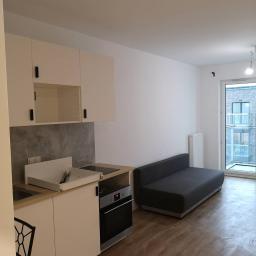 Venkonsv - Nowoczesne wnętrze kawalerki z aneksem kuchennym w trakcie montażu, widoczna wysuwana szuflada z akcesoriami kuchennymi, białe szafki z czarnymi uchwytami, szara sofa i drewnopodobna podłoga.