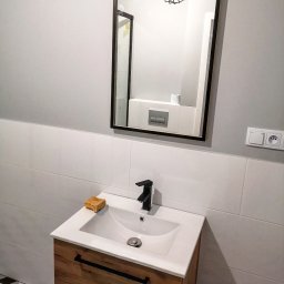 Venkonsv - Nowoczesna łazienka z umywalką nablatową na drewnianej szafce z czarnymi uchwytami, czarną baterią i lustrem w czarnej ramie, na blacie leży gąbka, widoczna podłoga w geometryczny wzór.