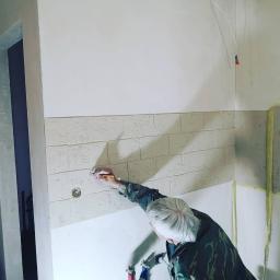 Venkonsv - Pracownik w wojskowej kurtce nakłada gładź imitującą cegłę na ścianę w łazience, widoczne instalacje hydrauliczne i otwór pod gniazdko.