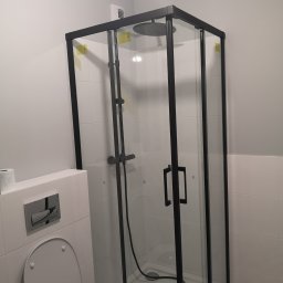 Venkonsv - Nowoczesna kabina prysznicowa z czarnymi profilami w wykończonej łazience, widoczna toaleta z ukrytą spłuczką i fragment podłogi w geometryczny wzór.