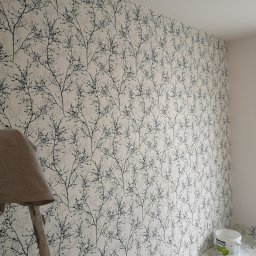 Venkonsv - Ściana pokryta tapetą w delikatny wzór czarnych gałązek, widoczna drabina malarska i wiadro z farbą, wskazujące na prace wykończeniowe w trakcie realizacji.