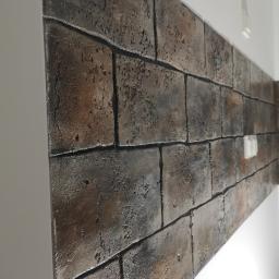 Venkonsv - Dekoracyjna ściana imitująca mur z cegły, wykonana z tynku gipsowego z widoczną fakturą i ciemnymi spoinami, oświetlona z góry, z widocznym fragmentem białej ściany obok.