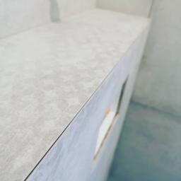 Venkonsv - Szara, teksturowana płyta ceramiczna imitująca beton, ułożona na półce w łazience, widoczna z bliska z perspektywy lekko od dołu, z fragmentem ściany i podłogi w tle.