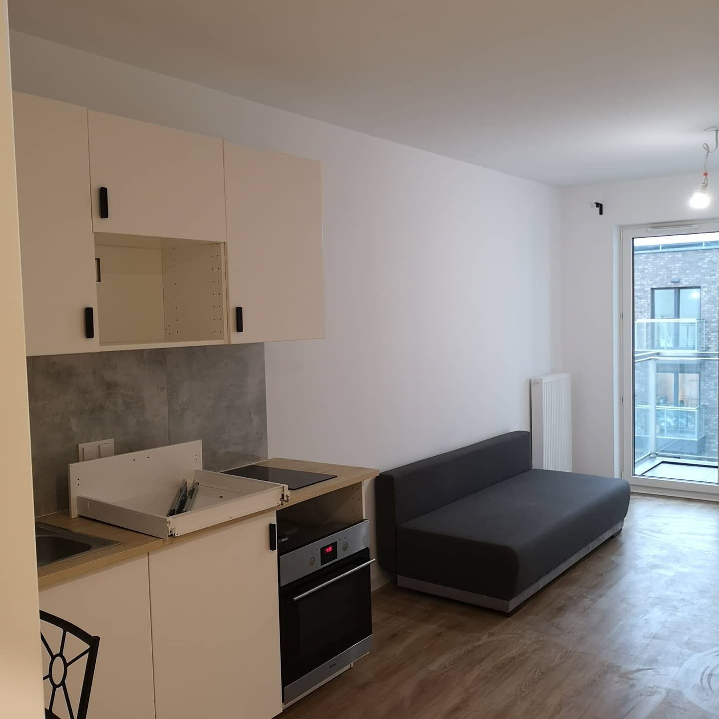 Nowoczesne wnętrze kawalerki z aneksem kuchennym w trakcie montażu, widoczna wysuwana szuflada z akcesoriami kuchennymi, białe szafki z czarnymi uchwytami, szara sofa i drewnopodobna podłoga.