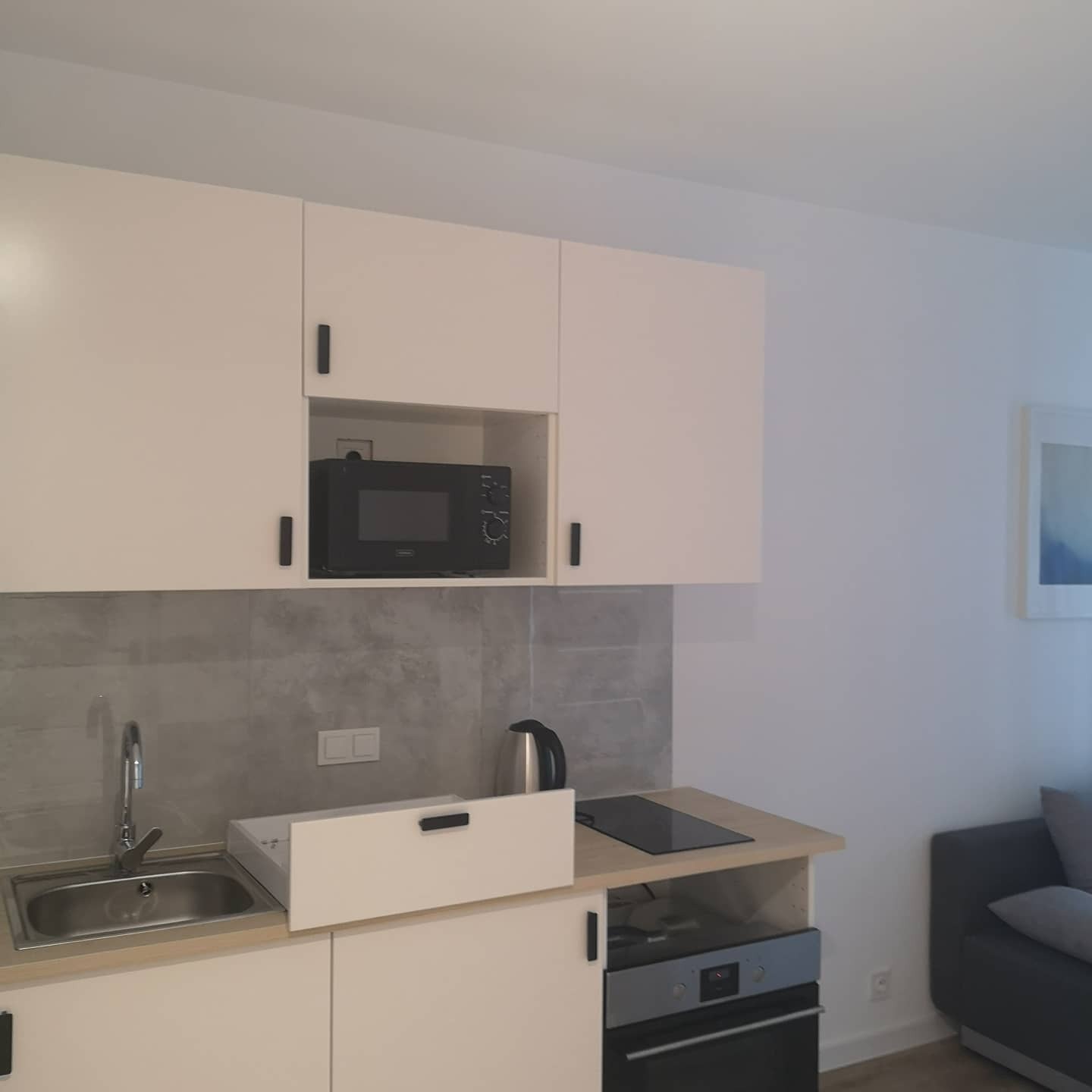Jasna, minimalistyczna kuchnia z beżowymi szafkami, szarymi płytkami nad blatem, czarną mikrofalówką i płytą indukcyjną, z widocznym fragmentem szarej sofy w tle.