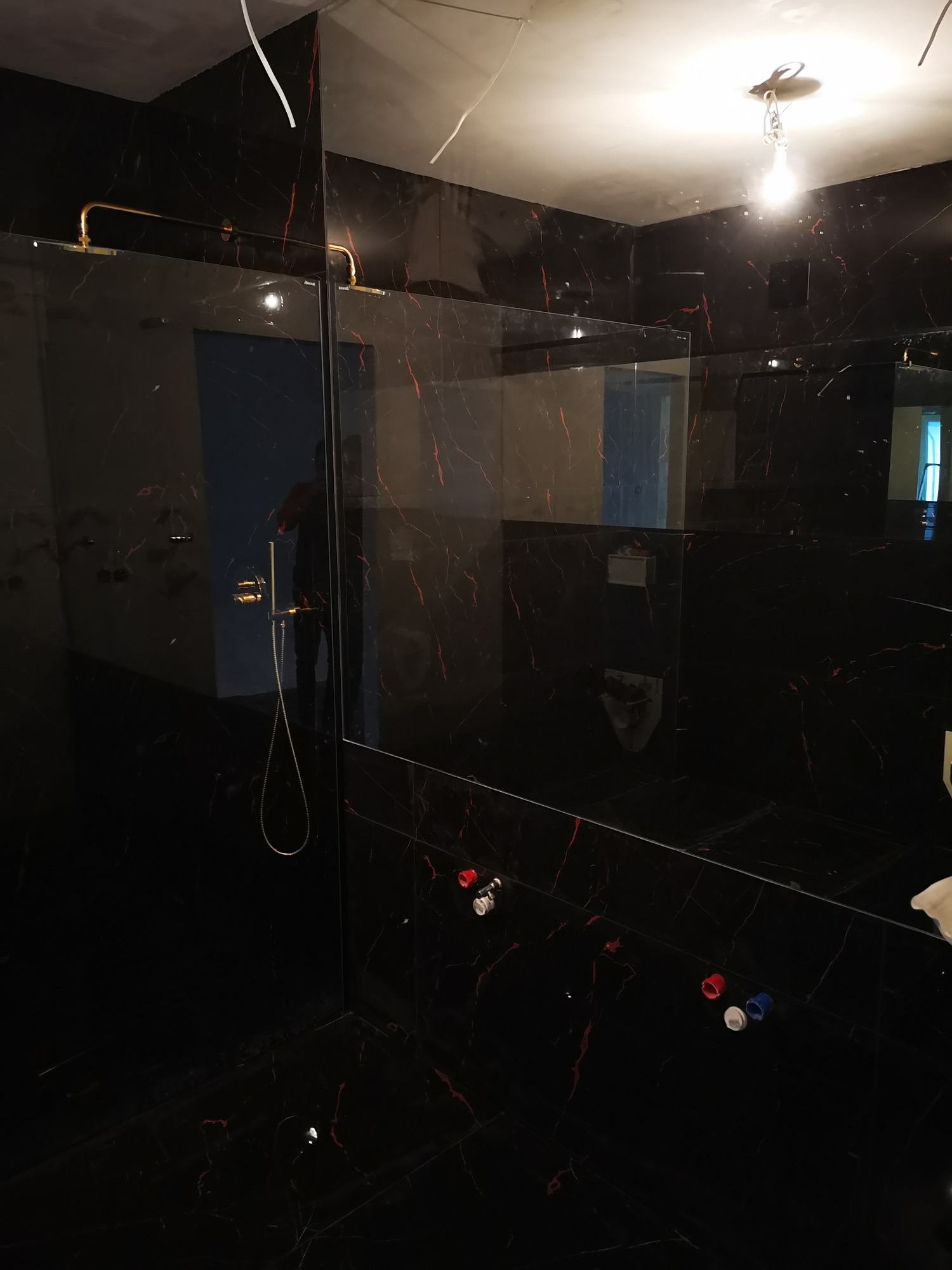 Czarne płytki z czerwonymi żyłkami w wykończonej łazience z zamontowaną szklaną kabiną prysznicową i złotą armaturą, widoczne niezabezpieczone końcówki przewodów elektrycznych pod sufitem.