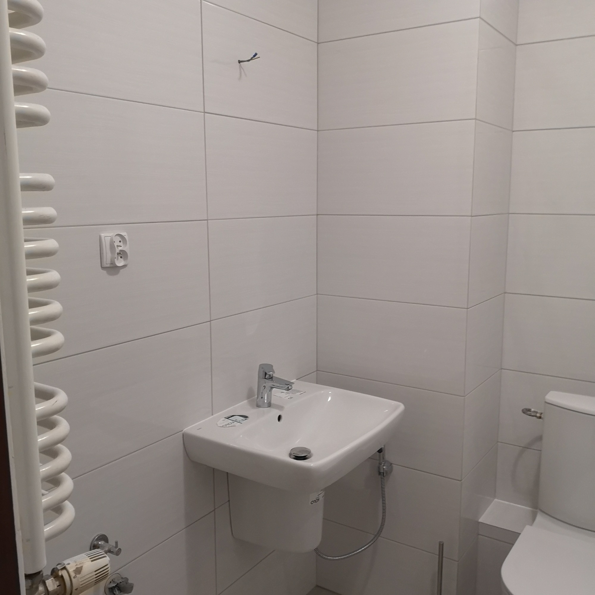 Biała umywalka podwieszana z chromowaną baterią i odpływem, obok toaleta i grzejnik drabinkowy, całość w łazience wyłożonej białymi płytkami z szarą fugą. Widoczny wystający kabel ze ściany.