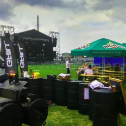 Plenerowa impreza z cateringiem: scena muzyczna w tle, rzędy stolik&oacute;w pod parasolami z logo piwa, stanowisko z grillem i beczkami.