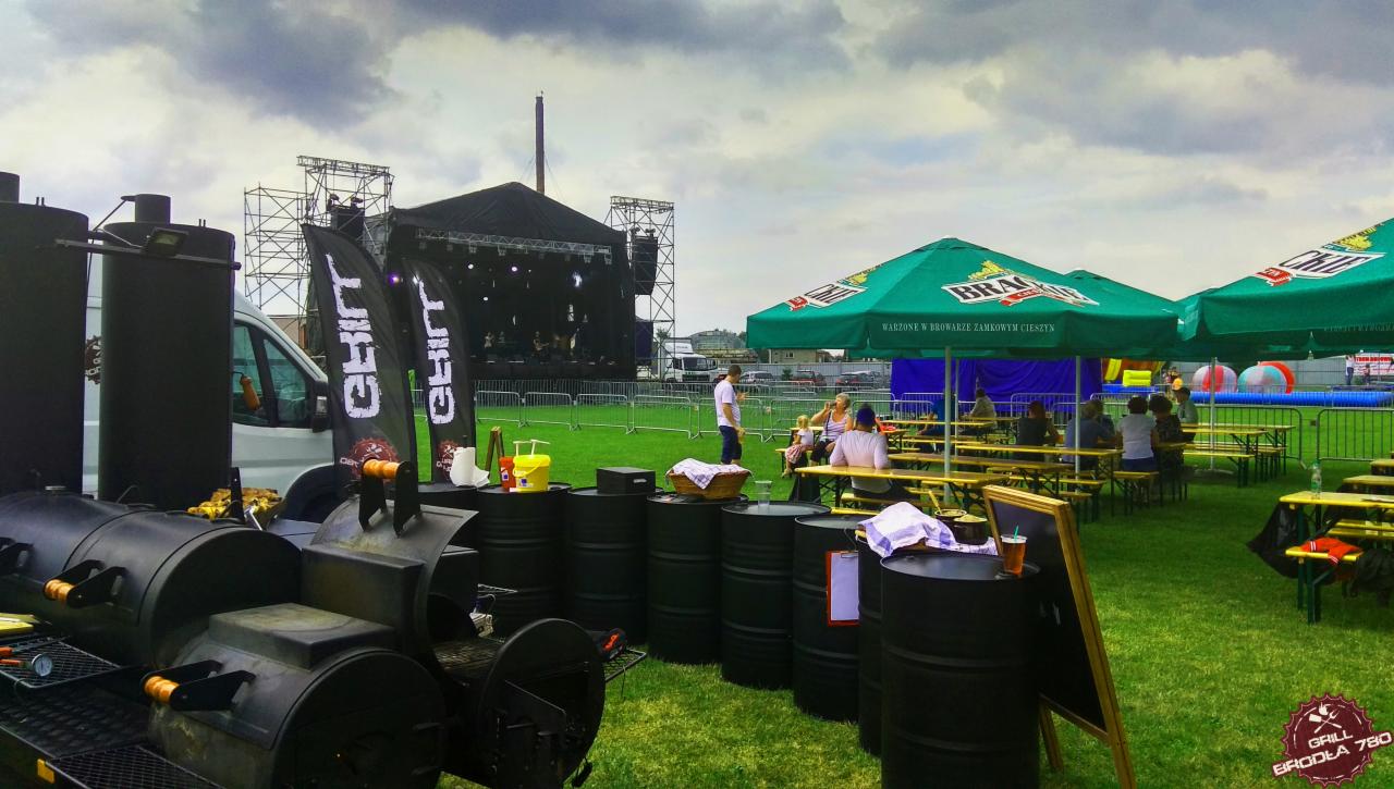 Plenerowa impreza z cateringiem: scena muzyczna w tle, rzędy stolików pod parasolami z logo piwa, stanowisko z grillem i beczkami.