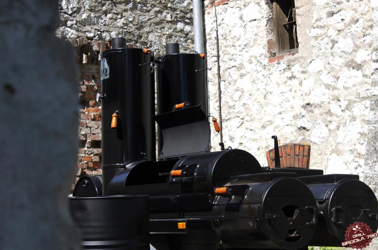 Duży, czarny grill smoker z pomarańczowymi uchwytami na tle kamiennej ściany i częściowo zasłonięty fragmentem muru.