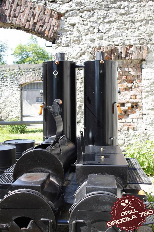 Duży, czarny grill BBQ z dwoma pionowymi kominami i logiem 'Grill Broda 780' na tle kamiennego muru w Czernichowie.