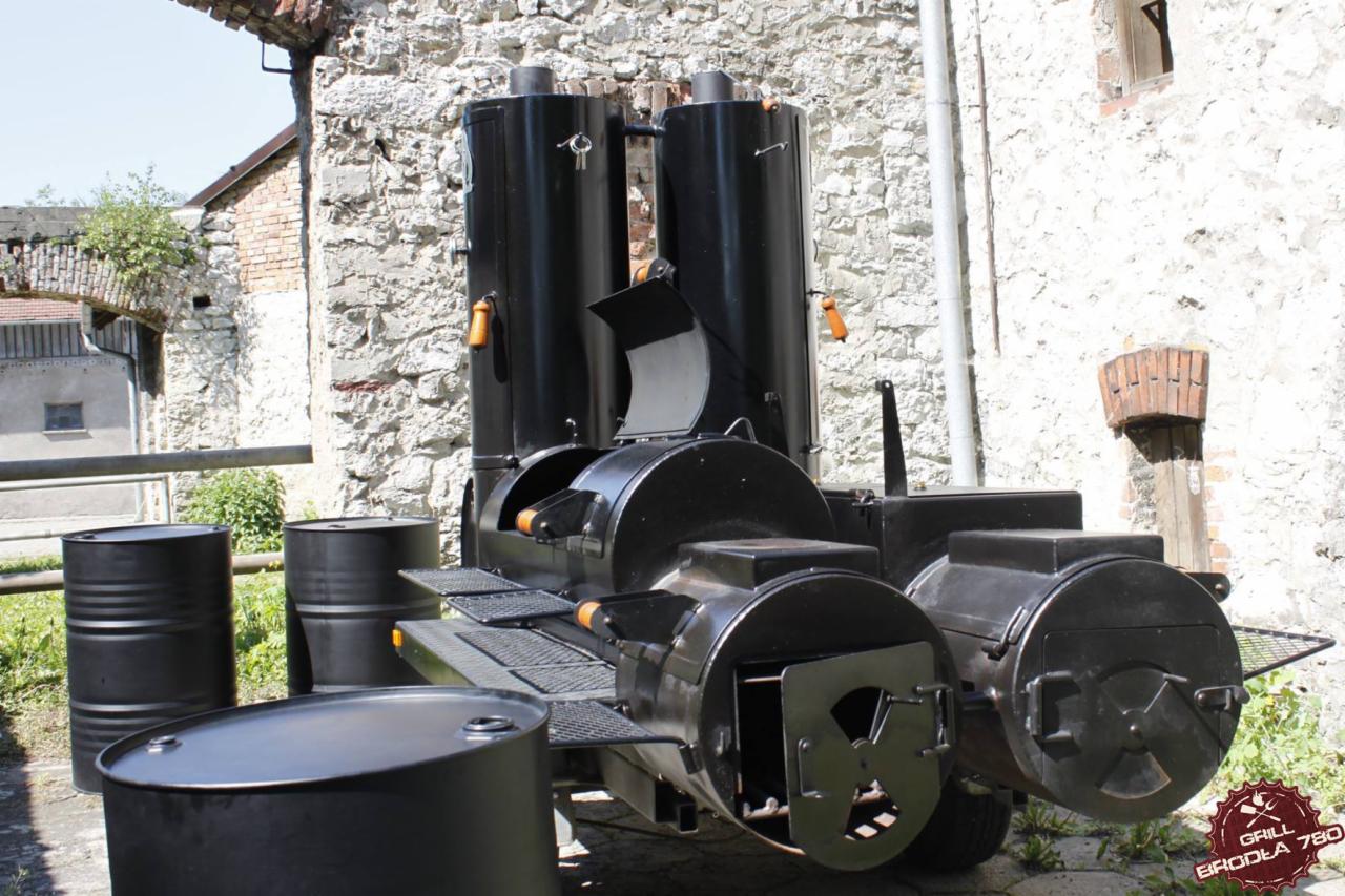 Duży, czarny grill smoker z dwoma kominami i trzema beczkami, ustawiony na zewnątrz na tle kamiennej ściany i budynków.