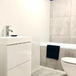 Nowoczesna łazienka z białą umywalką na szafce z połyskiem, chromowaną baterią, szarą płytką imitującą beton, ciemnoniebieskim pasem na suficie, dywanikiem i ręcznikiem.