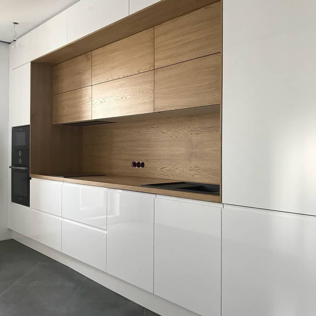 Minimalistyczna kuchnia z białymi, błyszczącymi frontami szafek dolnych i drewnianymi szafkami górnymi oraz panelem ściennym z widocznymi gniazdkami, piekarnik w zabudowie po lewej stronie.