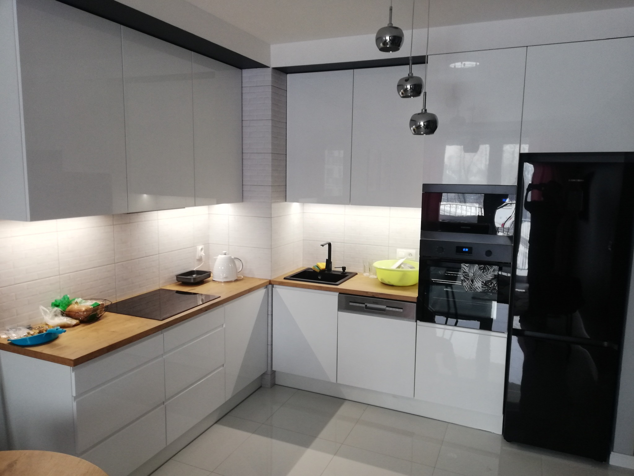 Nowoczesna kuchnia z białymi, lakierowanymi szafkami, drewnianym blatem i czarną lodówką, oświetlona trzema wiszącymi lampami.