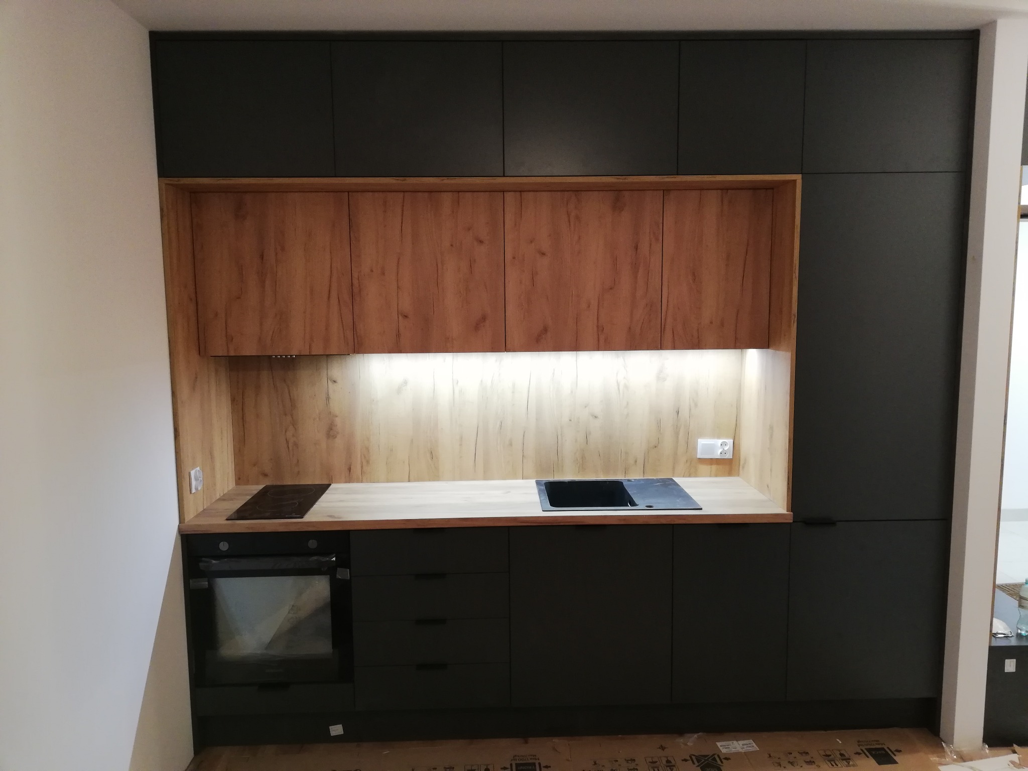 Nowoczesna zabudowa kuchenna w Warszawie: grafitowe szafki górne i dolne kontrastują z drewnopodobnym wykończeniem ściany i szafek wiszących. Minimalistyczny design z oświetleniem LED podszafkowym.