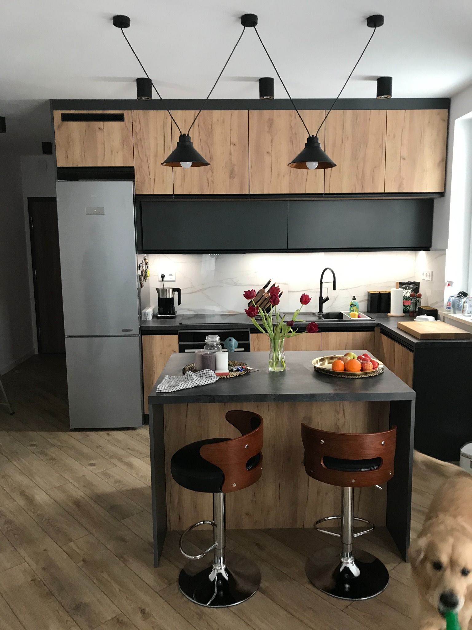 Nowoczesna kuchnia z drewnopodobnymi frontami, czarnymi detalami i wyspą z dwoma hokerami; na blacie wazon z tulipanami i misa z owocami. Pies w prawym dolnym rogu.