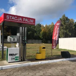 Stacja paliw ZMG z dystrybutorami, koszem na śmieci i flagami reklamowymi, widok w słoneczny dzień.