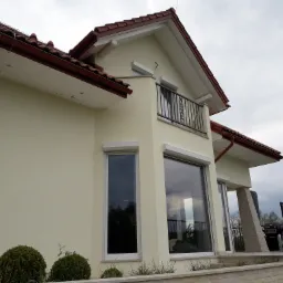 Jasna, otynkowana elewacja domu z balkonem, widoczne okna z roletami, brązowa dachówka i fragment kamiennej podmurówki.