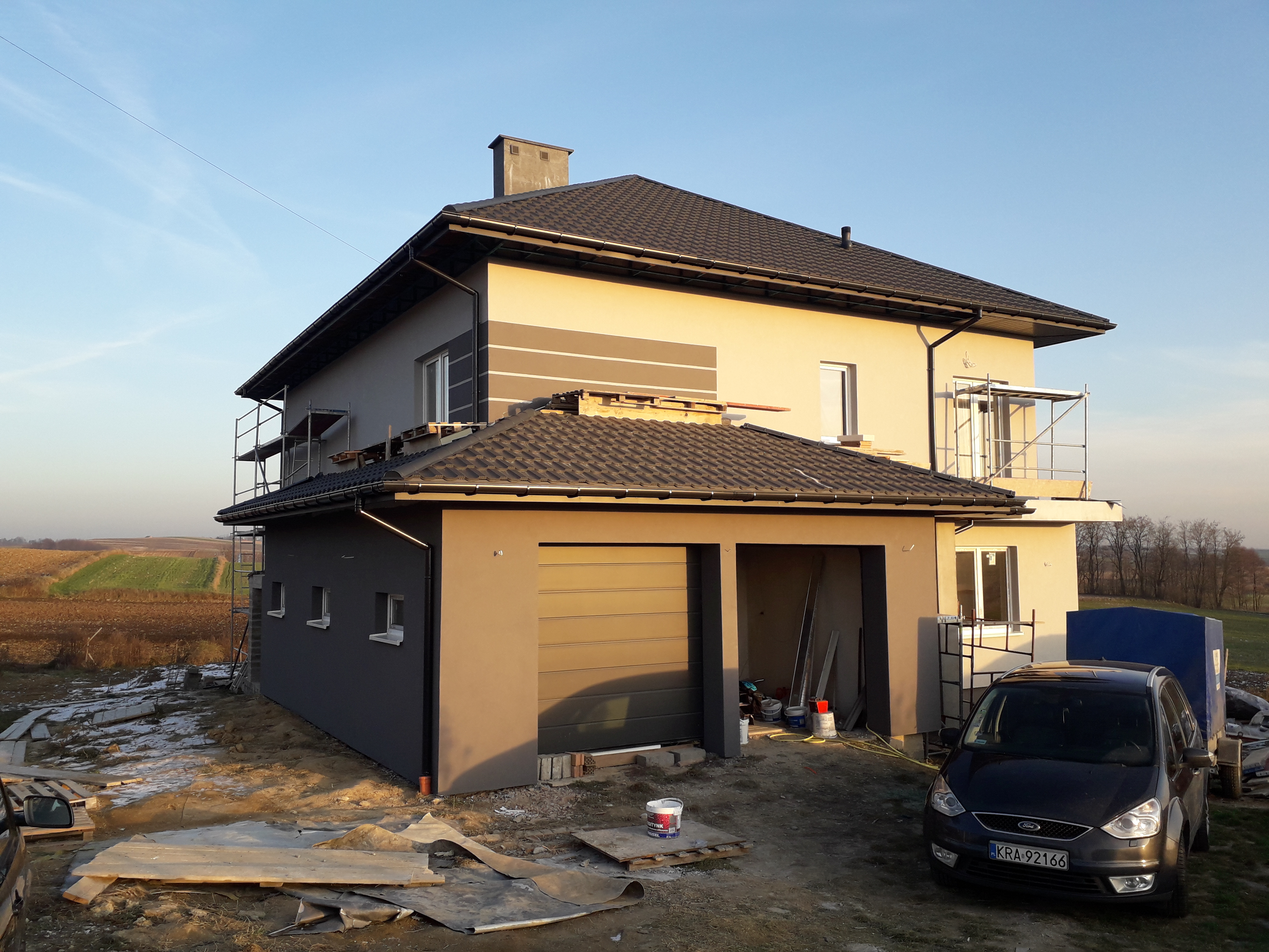 Dwukondygnacyjny dom z garażem w trakcie wykańczania elewacji, widoczne rusztowania, częściowo pomalowana fasada w odcieniach szarości i beżu, na pierwszym planie samochód osobowy i wiadro z farbą.