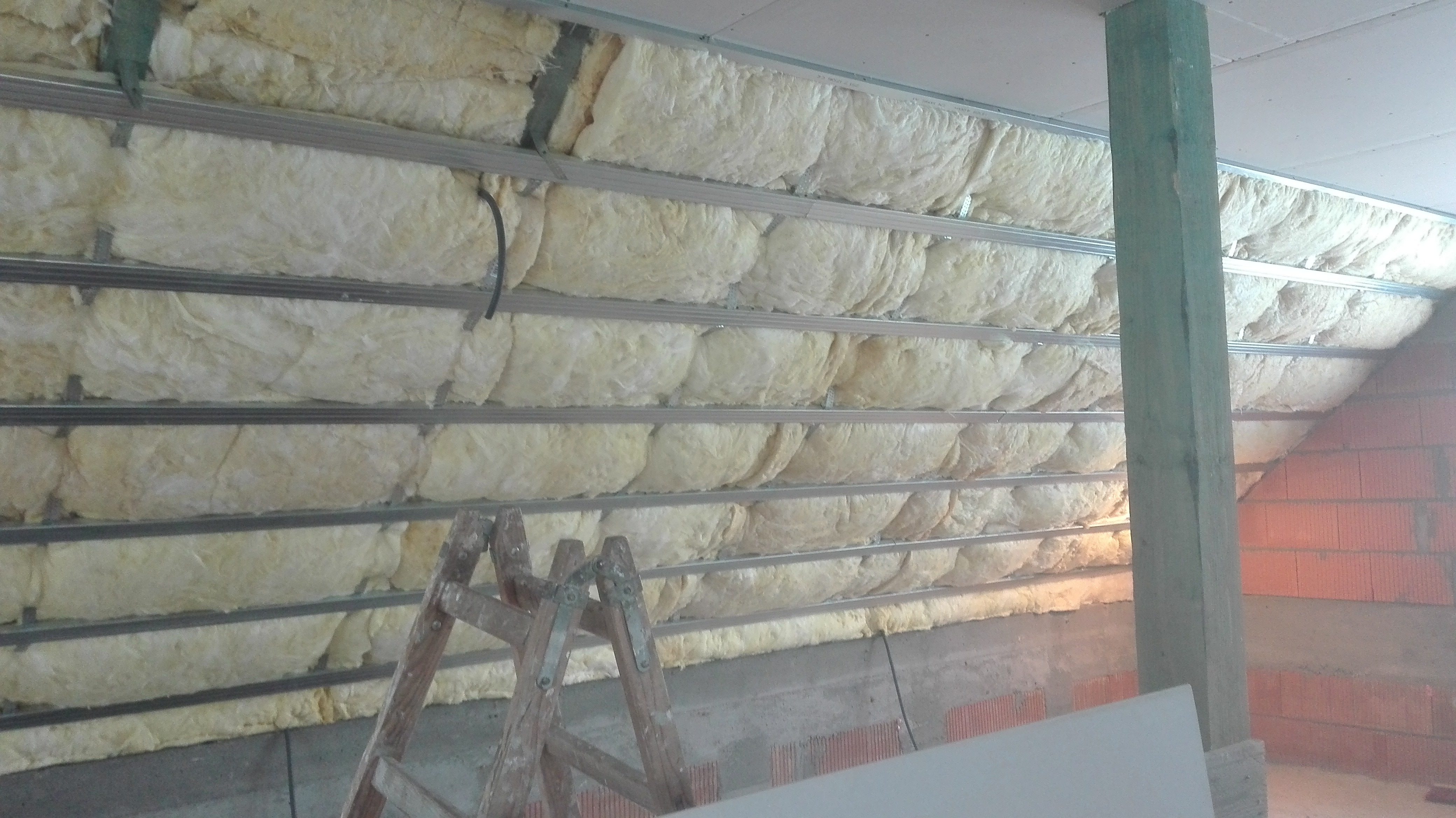 Montaż izolacji z wełny mineralnej w poddaszu, widoczne metalowe profile, częściowo otynkowana ściana z czerwonej cegły i drabina malarska.