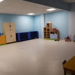 Jasne wnętrze sali zabaw z błękitnymi ścianami, wyposażonej w różnokolorowe szafki, regały z zabawkami i leżanki dla dzieci.