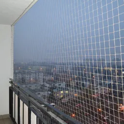 Zabezpieczony siatką balkon z widokiem na miasto o zmierzchu, metalowa balustrada w dolnej części kadru.