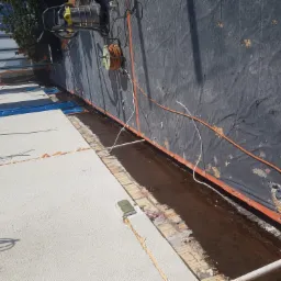 Przygotowanie do naprawy rynny dachowej: widoczna oczyszczona przestrzeń, narzędzia malarskie i kable na tle pokrycia dachowego i elewacji budynku.