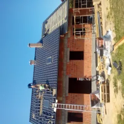 Montaż czarnej dachówki na nowym domu z cegły, widoczni dekarze, częściowo ukończona połać dachu z oknami dachowymi, drabina oparta o budynek, błękitne niebo.