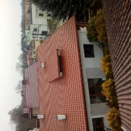 Nowy, czerwony dach z dachówki ceramicznej, widok z góry na budynek w trakcie wykańczania elewacji, z widocznymi oknami i otoczeniem.