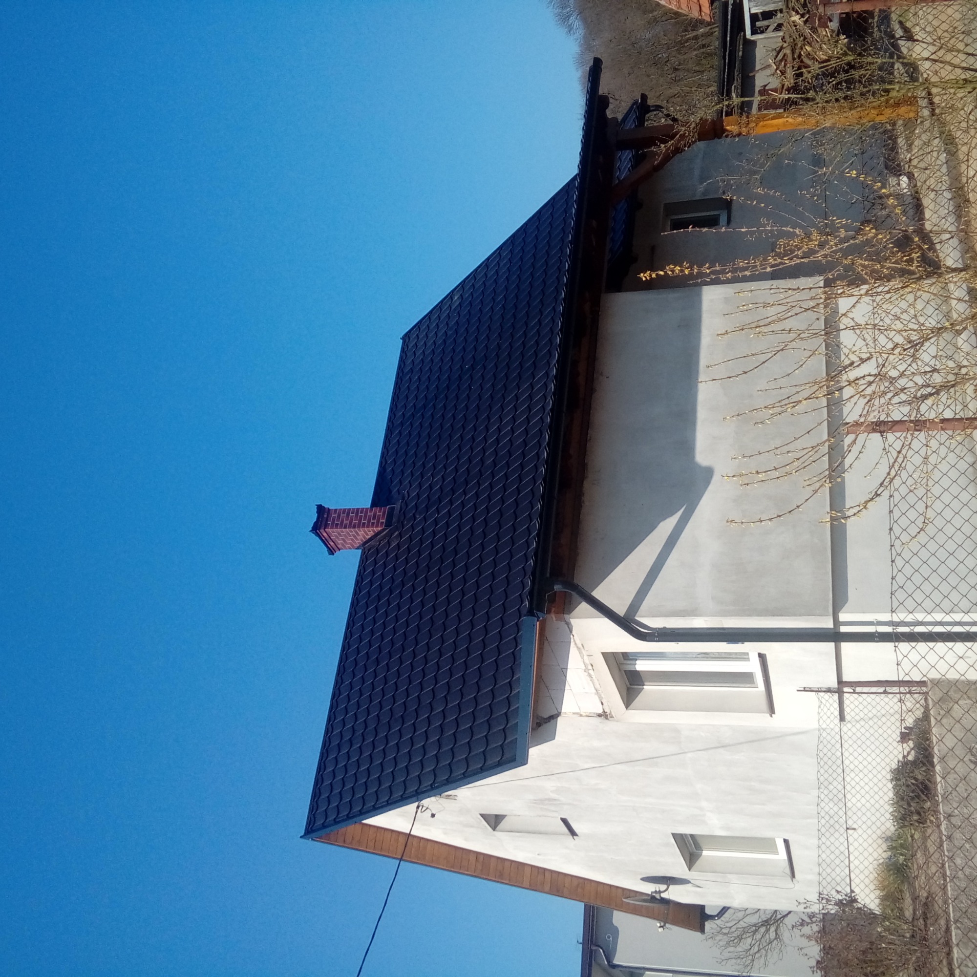 Ciemny, blaszany dach z ceglanym kominem na tle błękitnego nieba, widoczny fragment elewacji budynku z oknami i rynnami.