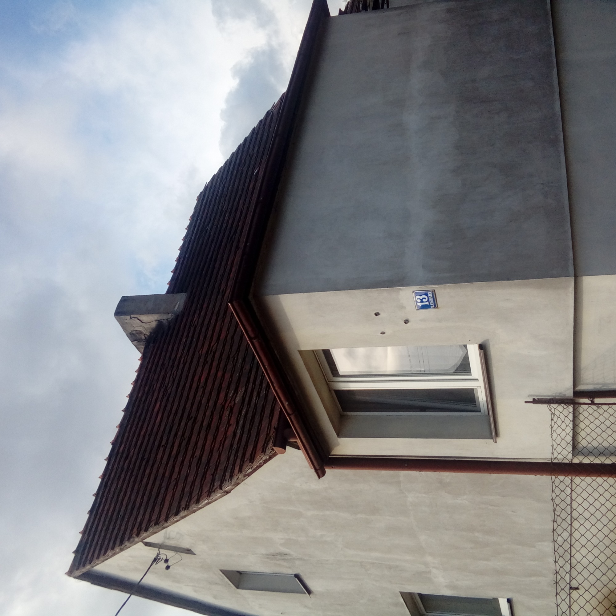 Fragment domu z widoczną częścią brązowego dachu, ścianą z numerem 13 i oknem, sfotografowany pod kątem z dołu.